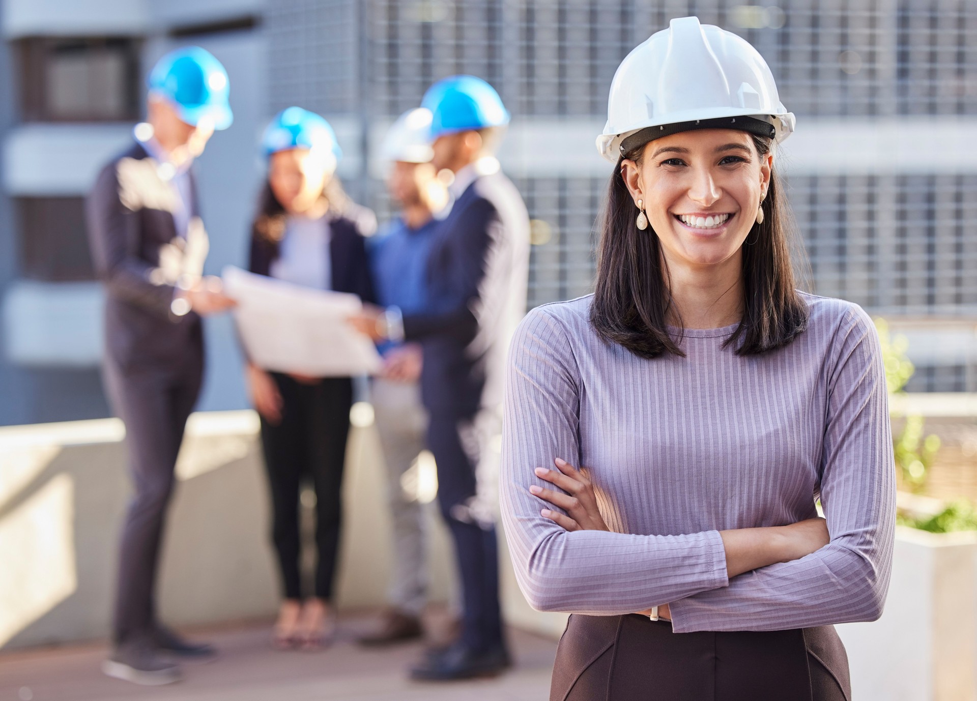 Shot of a young businesswoman standing with her arms folded and wearing a hardhat while her colleagues stand behind her